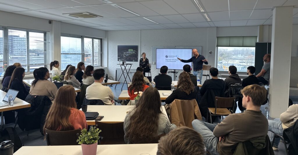 Een volle collegezaal waar studenten aandachtig luisteren naar twee gastsprekers; één persoon staat voor een scherm te presenteren terwijl de ander naast het scherm zit, in een lichte ruimte met grote ramen en uitzicht op de stad.