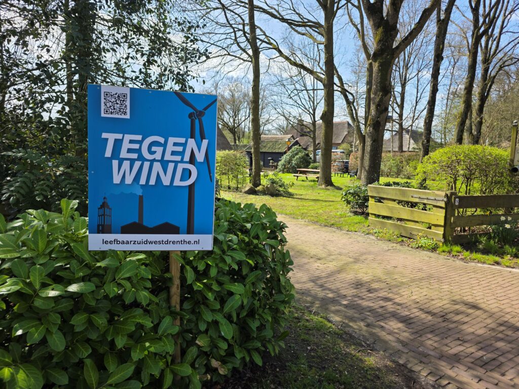 Een foto van een blauw bord met witte letters TEGEN WIND in een groene omgeving.